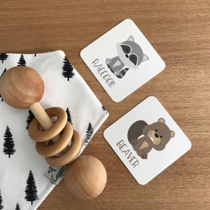 May include: A wooden baby rattle with a brown and white design. The rattle is on a white and black fabric with a tree pattern. There are two cards with animal illustrations and the words "Raccoon" and "Beaver".