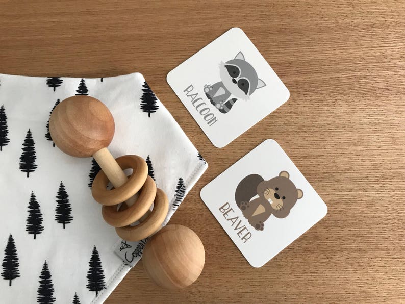 May include: A wooden baby rattle with a brown and white design. The rattle is on a white and black fabric with a tree pattern. There are two cards with animal illustrations and the words "Raccoon" and "Beaver".