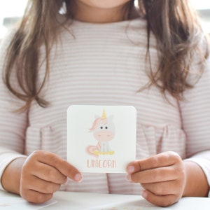 May include: A child holds a pink and white card with a cartoon unicorn illustration and the word "unicorn" printed below. The card is part of a memory game set from Goodnight Fox.