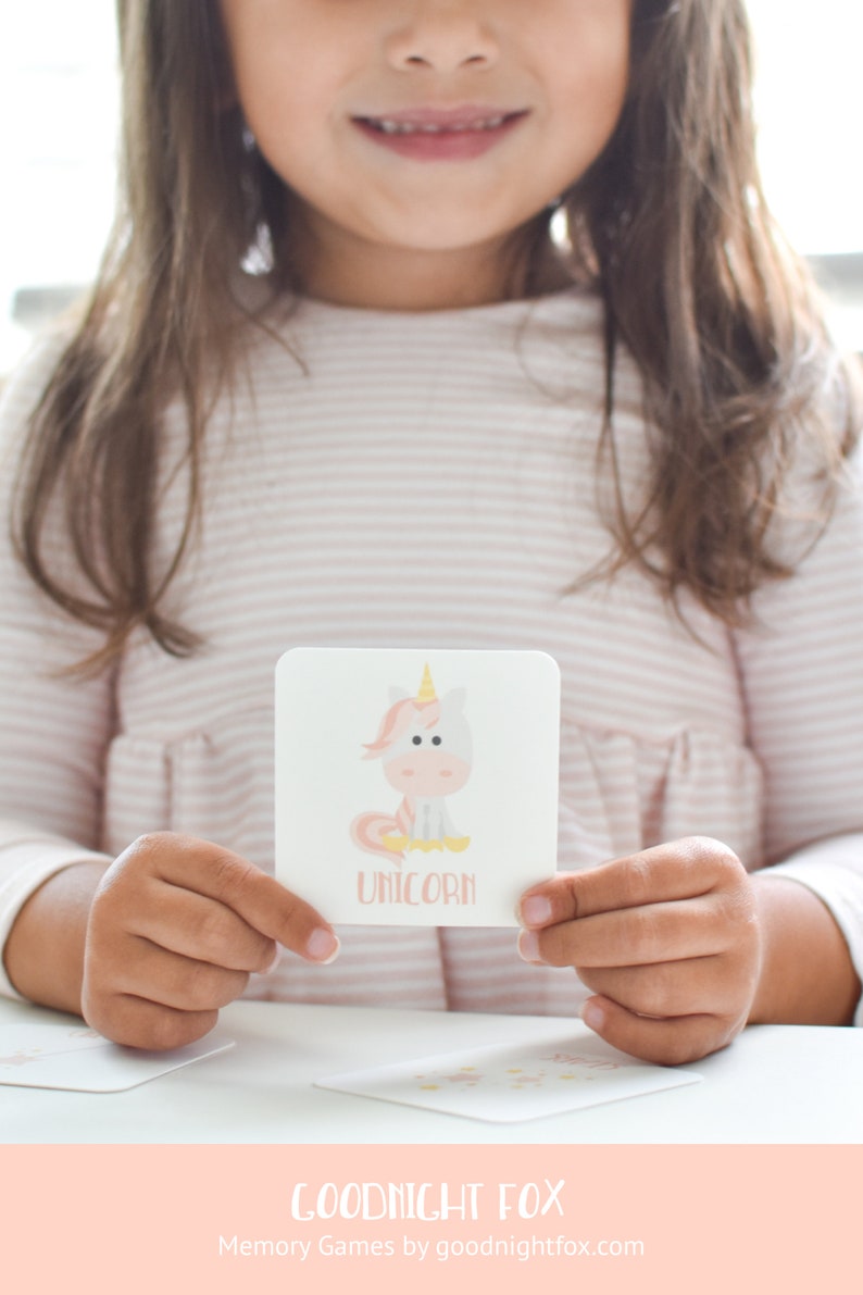 May include: A child holds a pink and white card with a cartoon unicorn illustration and the word "unicorn" printed below. The card is part of a memory game set from Goodnight Fox.