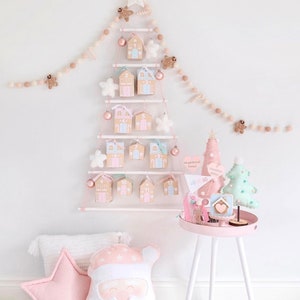 Puede incluir: Un calendario de adviento de árbol de Navidad rosa y blanco con casas de jengibre y una estrella en la parte superior. El calendario está colgado en una pared blanca con una guirnalda rosa y blanca. Hay una almohada de Papá Noel rosa y blanca y una mesa auxiliar rosa con una casa de jengibre y un árbol de Navidad.