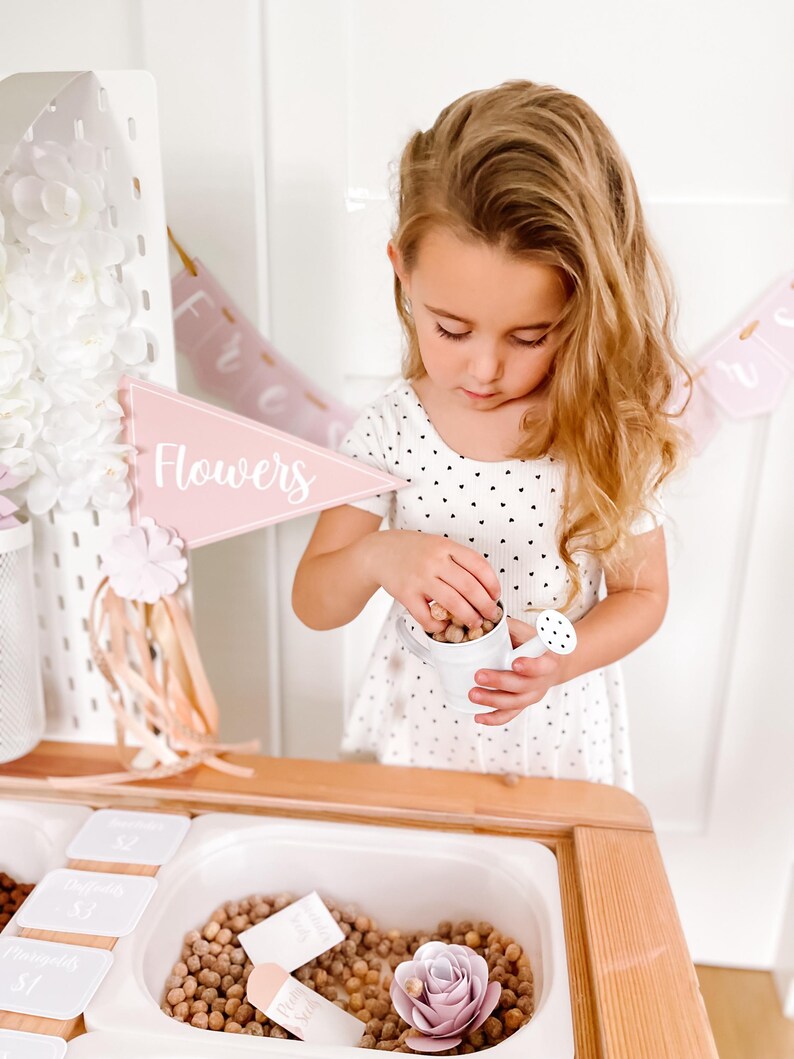 May include: A child in a white dress with black heart patterns holds a small white watering can. A pink banner with the word "Flowers" is visible. The scene includes a wooden table with a white bin filled with brown objects.