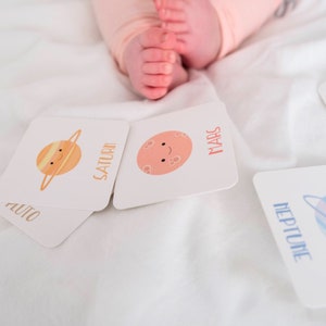 May include: A set of colorful flashcards featuring illustrations of planets with names like Saturn, Mars, Uranus, Neptune, and Pluto. The cards are on a white surface.