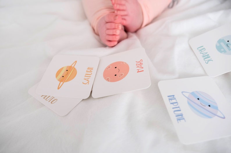 May include: A set of colorful flashcards featuring illustrations of planets with names like Saturn, Mars, Uranus, Neptune, and Pluto. The cards are on a white surface.