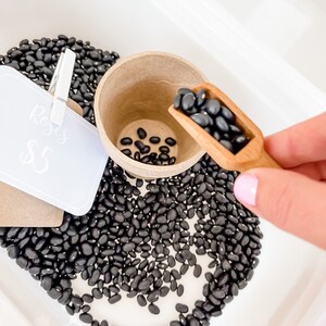 May include: A white tray filled with black beans, a small paper cup, and a wooden scoop. A sign clipped to the tray reads "Roses" and "$5". The beans are being scooped into the cup.