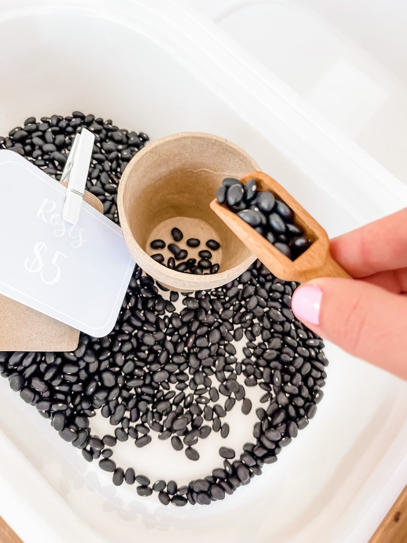 May include: A white tray filled with black beans, a small paper cup, and a wooden scoop. A sign clipped to the tray reads "Roses" and "$5". The beans are being scooped into the cup.