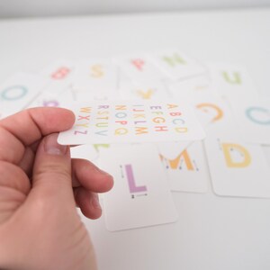 May include: A white card with the alphabet printed in rainbow colors. The letters are arranged in alphabetical order. The card is being held by a hand.