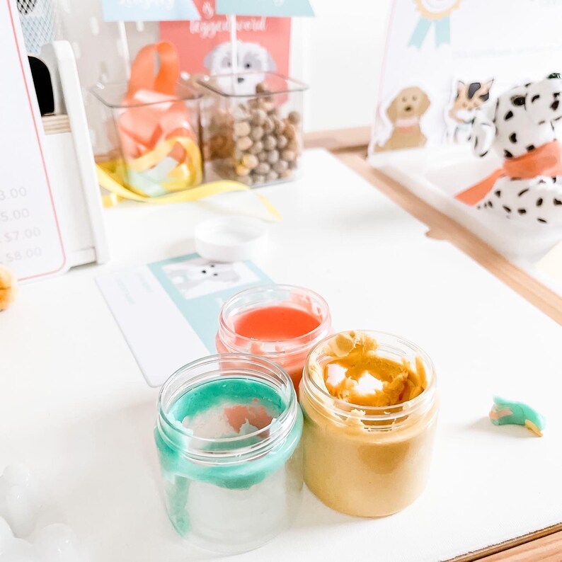 May include: Three small glass jars with lids containing different colored play dough. The jars are on a white surface. The play dough is teal, orange, and yellow.