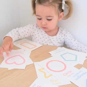 K&ouml;nnte beinhalten: Ein Kind lernt Formen mit bunten Lernkarten. Die Karten zeigen ein Herz, ein Quadrat, einen Stern, einen Kreis und ein Sechseck mit dem Namen jeder Form in Englisch und Spanisch.