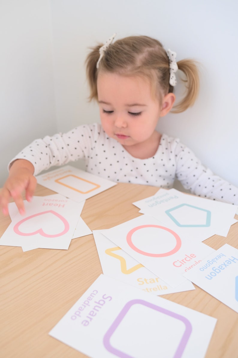 K&ouml;nnte beinhalten: Ein Kind lernt Formen mit bunten Lernkarten. Die Karten zeigen ein Herz, ein Quadrat, einen Stern, einen Kreis und ein Sechseck mit dem Namen jeder Form in Englisch und Spanisch.