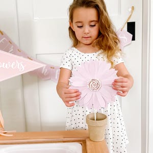 May include: A young girl holds a large, light pink paper flower with a detailed centre. The flower is in a small, brown paper pot. The girl wears a white dress with black heart-shaped patterns.