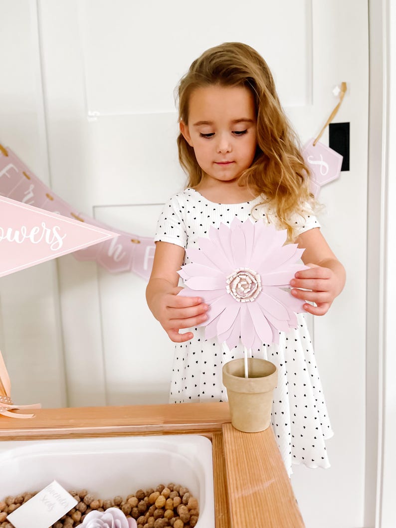 May include: A young girl holds a large, light pink paper flower with a detailed centre. The flower is in a small, brown paper pot. The girl wears a white dress with black heart-shaped patterns.