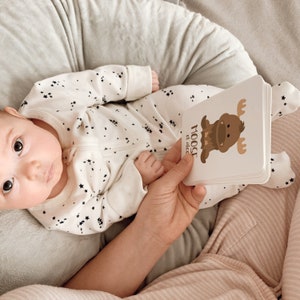 May include: A baby wearing a white onesie with black stars is holding a board book with a brown moose illustration and the text "MOOSE el alce".