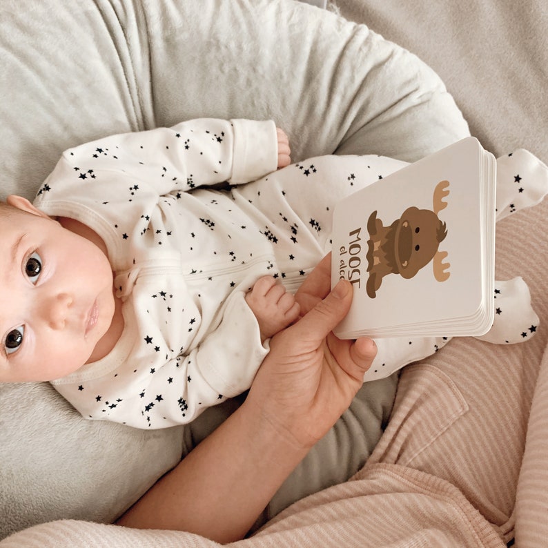 May include: A baby wearing a white onesie with black stars is holding a board book with a brown moose illustration and the text "MOOSE el alce".