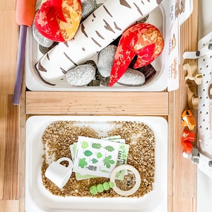 May include: A wooden play set with two white trays. The top tray has a birch log, red flames, and gray rocks. The bottom tray has a white cup, a magnifying glass, green beans, and a card with the text "Leaf Hunt".