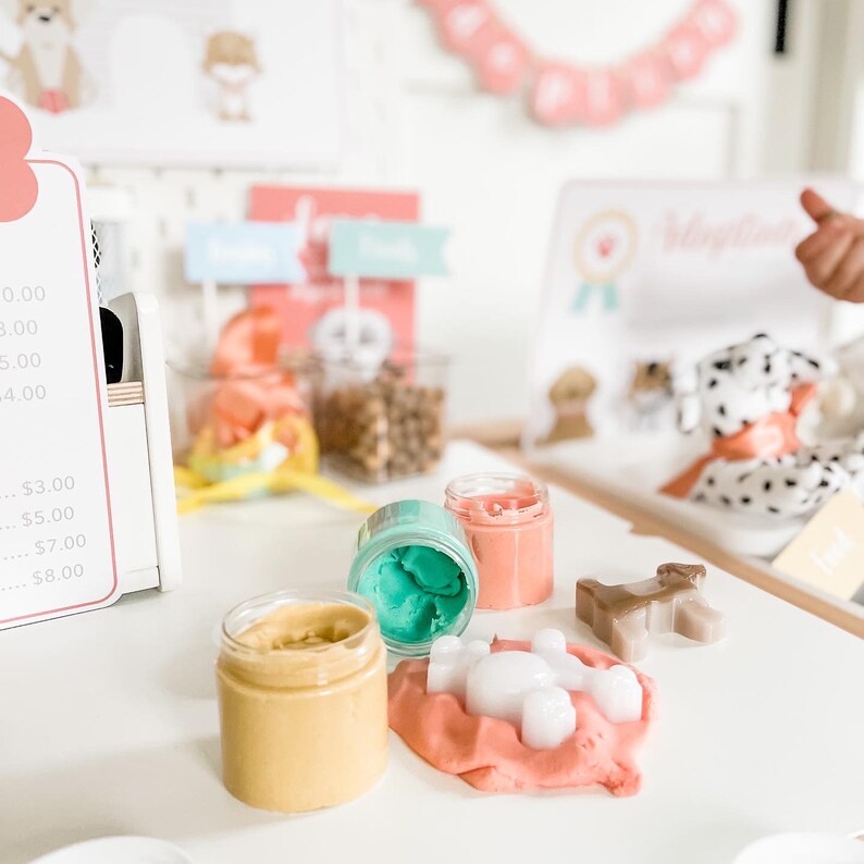 May include: A play dough set with three containers of play dough in pink, green, and yellow. The play dough is shaped into a dog and a bone. The containers are labeled with prices.