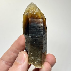 May include: A translucent, elongated quartz crystal point featuring a gradient of colours ranging from a pale, honey-yellow at the apex to a deep, smoky brown and dark charcoal grey toward the base, with visible horizontal striations along the sides.
