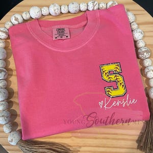May include: Pink softball t-shirt with a yellow softball with a red stitch design and the number 5 on it. The text "Kenslie" is written in white below the softball. The text "Young Southern Mess" is written in white at the bottom of the shirt.