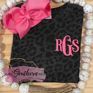 May include: A black and gray leopard print t-shirt with a pink monogram "RGS" on the front. The shirt is on a wooden surface with white beads.