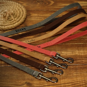 May include: A collection of personalized dog leashes in various colors, including gray, brown, tan, and pink. Each leash features a silver-colored clasp and is engraved with a dog's name. The leashes are arranged on a wooden surface.