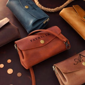 Op de afbeelding: Een assortiment leren portemonnees in verschillende kleuren, waaronder bruin, beige en blauw. Sommige portemonnees hebben gepersonaliseerde tekst zoals "Patricia" en "Catheryn". Elke portemonnee heeft een knoopsluiting en een riem of handvat.