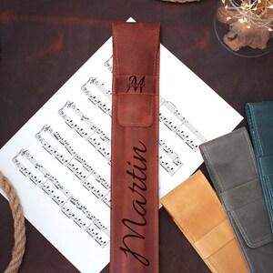 Op de afbeelding: Een bruine leren etui met de naam "Martin" erop gegraveerd. De etui ligt op een blad muziek met andere leren etuis in verschillende kleuren.