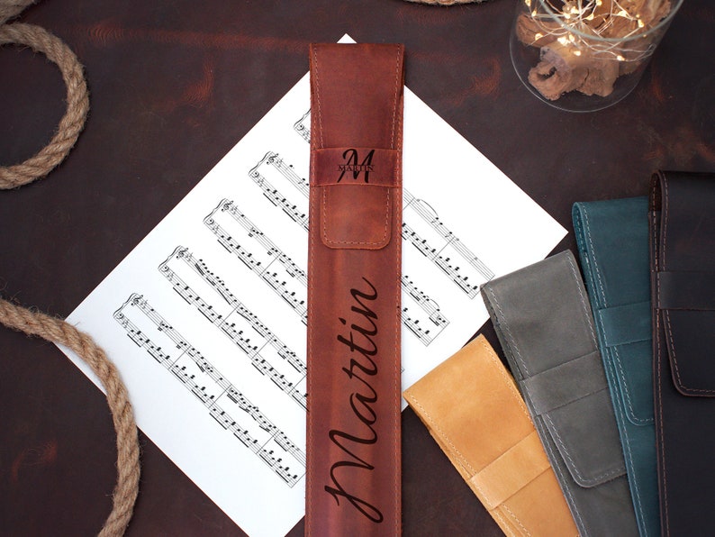 Op de afbeelding: Een bruine leren etui met de naam "Martin" erop gegraveerd. De etui ligt op een blad muziek met andere leren etuis in verschillende kleuren.