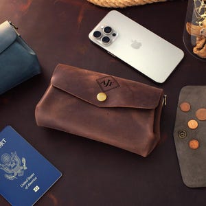 Op de afbeelding: Een verzameling lederwaren, waaronder een bruine leren etui met een gouden knoop en initialen, een blauwgroene etui en een grijze portemonnee. Een paspoort en een smartphone zijn ook aanwezig.