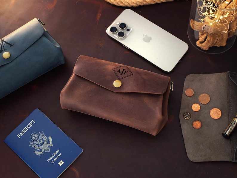 Op de afbeelding: Een verzameling lederwaren, waaronder een bruine leren etui met een gouden knoop en initialen, een blauwgroene etui en een grijze portemonnee. Een paspoort en een smartphone zijn ook aanwezig.
