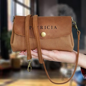 Op de afbeelding: Een bruine leren crossbodytas met de naam "PATRICIA" op de flap. De tas heeft een lange, verstelbare riem en een goudkleurige sluiting. De tas is ongeveer 20 cm breed.