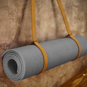 May include: A gray yoga mat with a brown leather strap hanging on a textured wall. The strap is looped around the mat and secured with a knot.