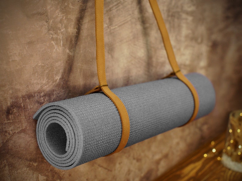 May include: A gray yoga mat with a brown leather strap hanging on a textured wall. The strap is looped around the mat and secured with a knot.