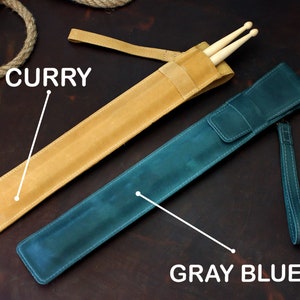 Op de afbeelding: Twee leren drumstick-etuis, een is currykleurig en de andere is grijsblauw. De curry-etui heeft een band en de grijsblauwe etui heeft een band en een polsband.