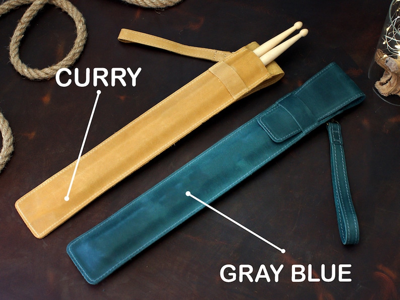Op de afbeelding: Twee leren drumstick-etuis, een is currykleurig en de andere is grijsblauw. De curry-etui heeft een band en de grijsblauwe etui heeft een band en een polsband.