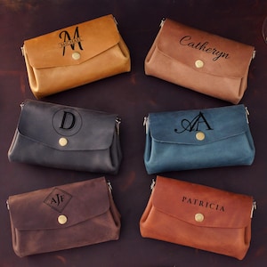 May include: Six leather crossbody bags in various colors: tan, brown, navy, blue, and rust. Each bag features a flap closure with a gold button and personalized text or initials. The bags are arranged on a dark brown surface.