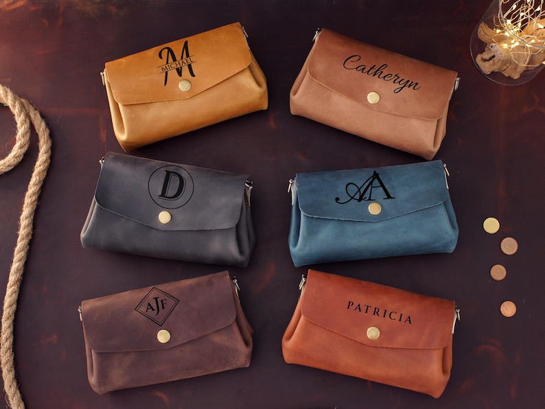 May include: Six leather crossbody bags in various colors: tan, brown, navy, blue, and rust. Each bag features a flap closure with a gold button and personalized text or initials. The bags are arranged on a dark brown surface.