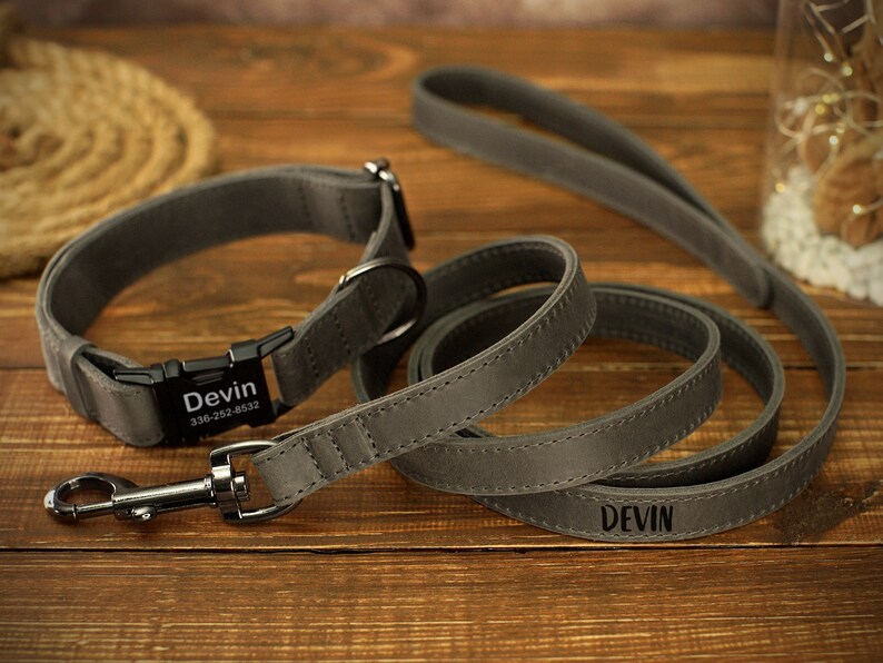 May include: A gray dog collar and leash set. The collar has a black buckle with the name "Devin" and a phone number. The matching leash also has the name "Devin" engraved. The set is made of a soft, durable material.