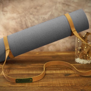 May include: A gray yoga mat with a brown leather strap and the name "Ariana" embossed on the strap.