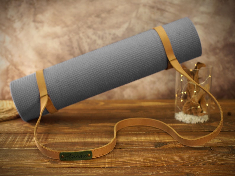 May include: A gray yoga mat with a brown leather strap and the name "Ariana" embossed on the strap.