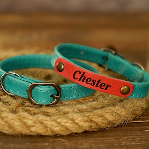 Puede incluir: Dos collares de cuero para gatos, uno azul turquesa y otro rojo con el nombre "Chester" grabado. Ambos collares tienen una peque&ntilde;a campana.