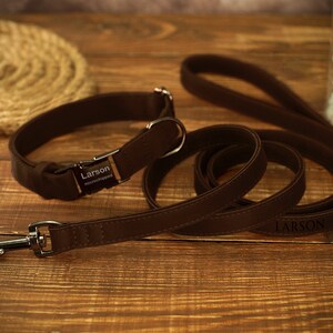 May include: A brown leather dog collar and matching leash set. The collar has a metal buckle and a nameplate that reads "Larson microchipped." The leash also has the name "Larson" printed on it. The set is displayed on a wooden surface.