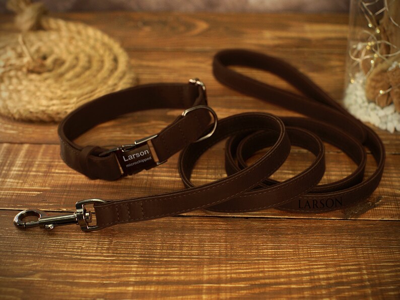 May include: A brown leather dog collar and matching leash set. The collar has a metal buckle and a nameplate that reads "Larson microchipped." The leash also has the name "Larson" printed on it. The set is displayed on a wooden surface.