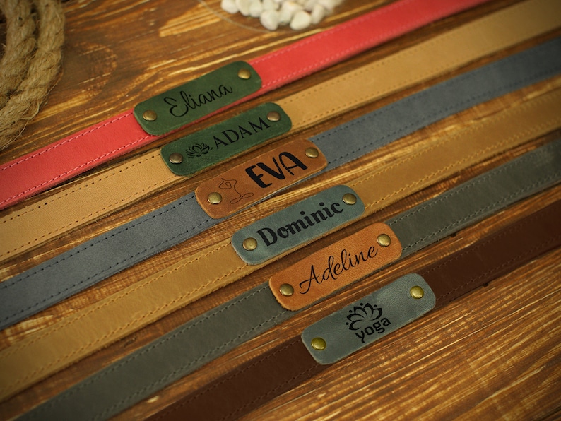 May include: A collection of six leather straps with personalized engraved name tags. The straps are in various colors, including red, brown, gray, and green. The name tags feature names like Eliana, Adam, Eva, Dominic, Adeline, and Yoga.