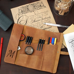 May include: A brown leather roll-up case with multiple pockets for pens, pencils, and other writing tools. The case has a personalized inscription that reads "M MICHAEL".