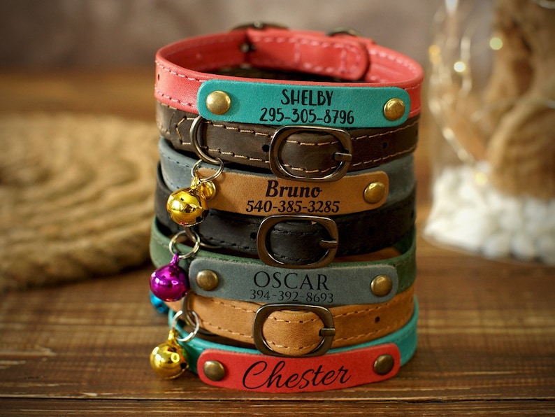 Puede incluir: Pila de cinco collares de cuero para gatos de colores con etiquetas de nombre personalizadas y n&uacute;meros de tel&eacute;fono. Los collares son marrones, grises, azul verdoso, beige y rosas. Cada collar tiene una campanilla dorada.