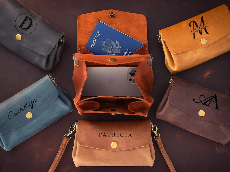 May include: A collection of leather crossbody bags in various colors, including brown, tan, blue, and gray. One bag is open, revealing a passport and a smartphone. Each bag features a personalized initial or name.
