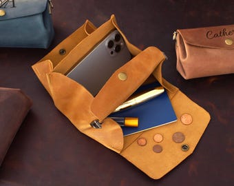 Könnte beinhalten: Eine offene, hellbraune Leder-Ablageschale mit Smartphone, Stift, kleinem Notizbuch und Münzen. Im Hintergrund befindet sich eine kleine braune Ledertasche. Die Schale wird mit einem Druckknopf verschlossen.