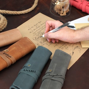 K&ouml;nnte beinhalten: Vier personalisierte Leder-Stiftrollen in Braun, Beige, Blau und Grau. Die Rollen sind aufgerollt und haben einen Riemen mit einem eingravierten Namen auf jeder. Die Namen sind Bessie, William, Anabel und Angelica.