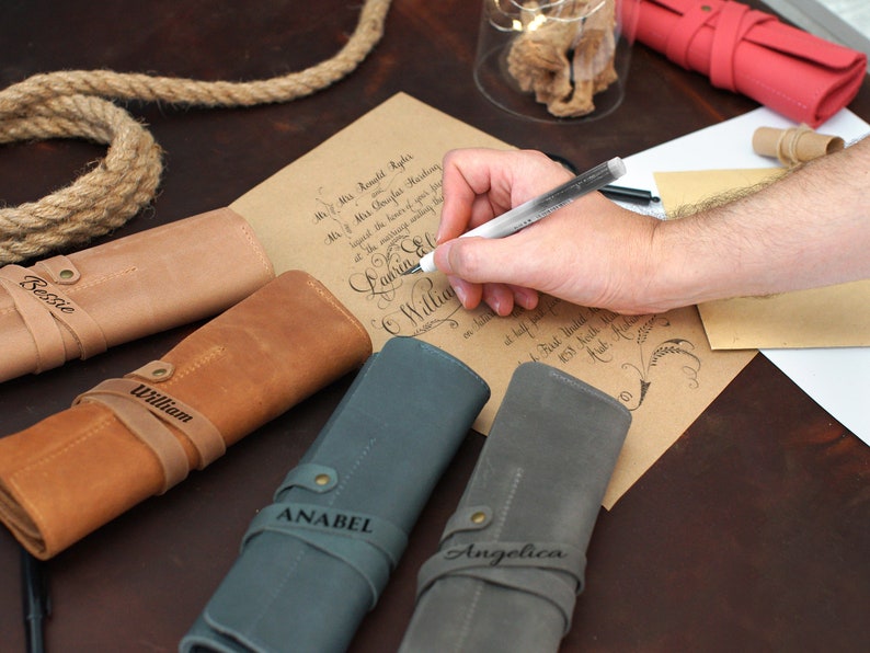 K&ouml;nnte beinhalten: Vier personalisierte Leder-Stiftrollen in Braun, Beige, Blau und Grau. Die Rollen sind aufgerollt und haben einen Riemen mit einem eingravierten Namen auf jeder. Die Namen sind Bessie, William, Anabel und Angelica.