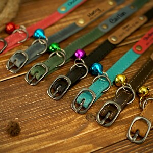 May include: A collection of personalized pet collars in various colors, including red, gray, green, black, teal, brown, and tan. Each collar features a metal buckle, a small bell, and a name tag. The collars are arranged on a wooden surface.